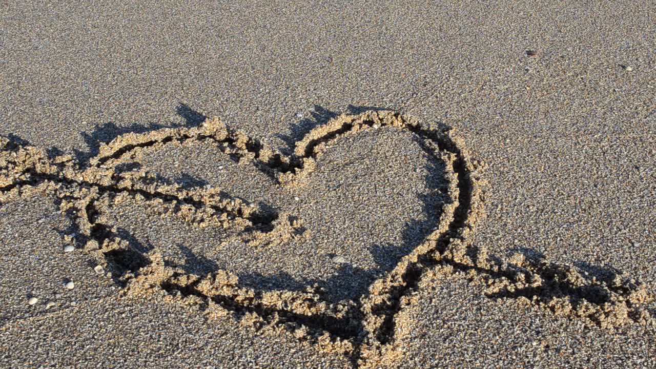 Un cuore disegnato sulla sabbia. Il cuore viene cancellato da un'onda marina. Riprese sulla spiaggia.