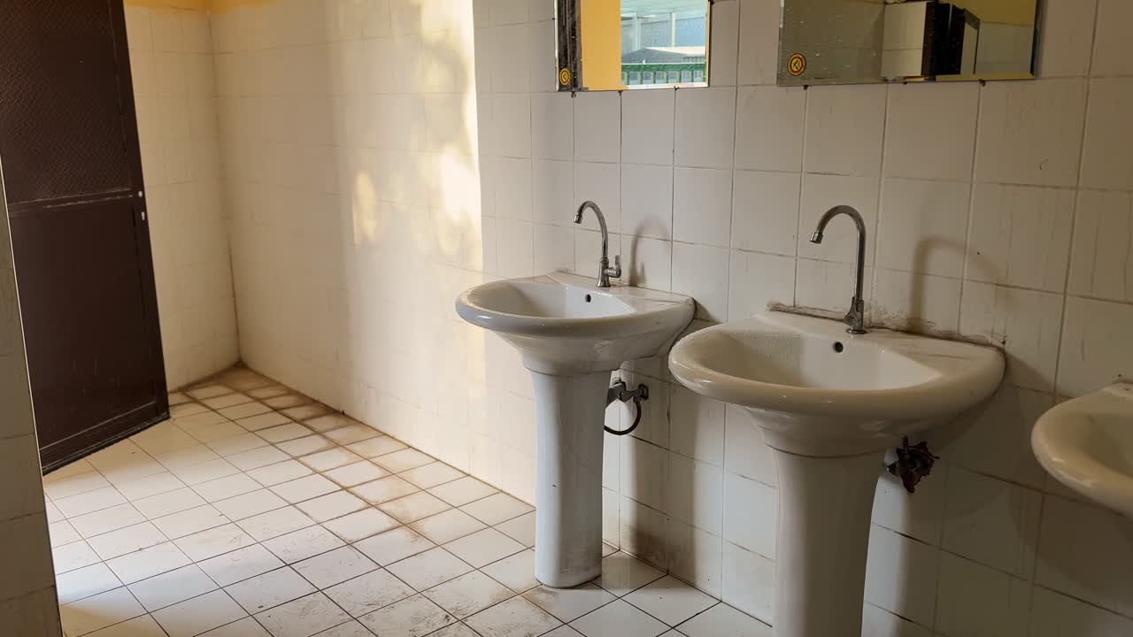 Vista interior amplia de un aseo público desgastado que presenta lavabos de pedestal, espejos de pared, superficies alicatadas y luz solar natural proyectando sombras por toda la zona de saneamiento.