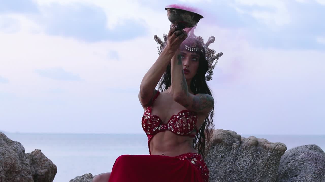 Beautiful, young belly dancer performing rhythmic moves by holding a colorful smoke bomb, on the beach. Slow motion