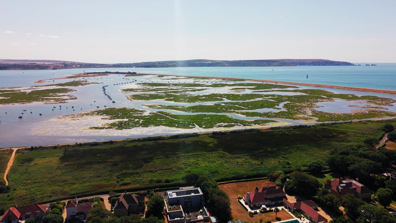 Riprese con drone sulla stessa palude salmastra e distese fangose di marea, barche ormeggiate e mare turchese verso un promontorio lontano, ora inquadrate per includere un villaggio sul lungomare con case dai tetti rossi e giardini