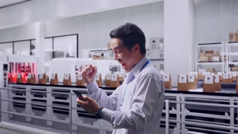side view of an asian male professional worker with his smartphone with conveyor for packaging coffee beans in bags at coffee factory, he raises his fist up with screaming goal