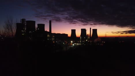 Illuminated-aerial-night-view-above-industrial-pipeline-chemical-factory-site-rising-over-tree-silhouette