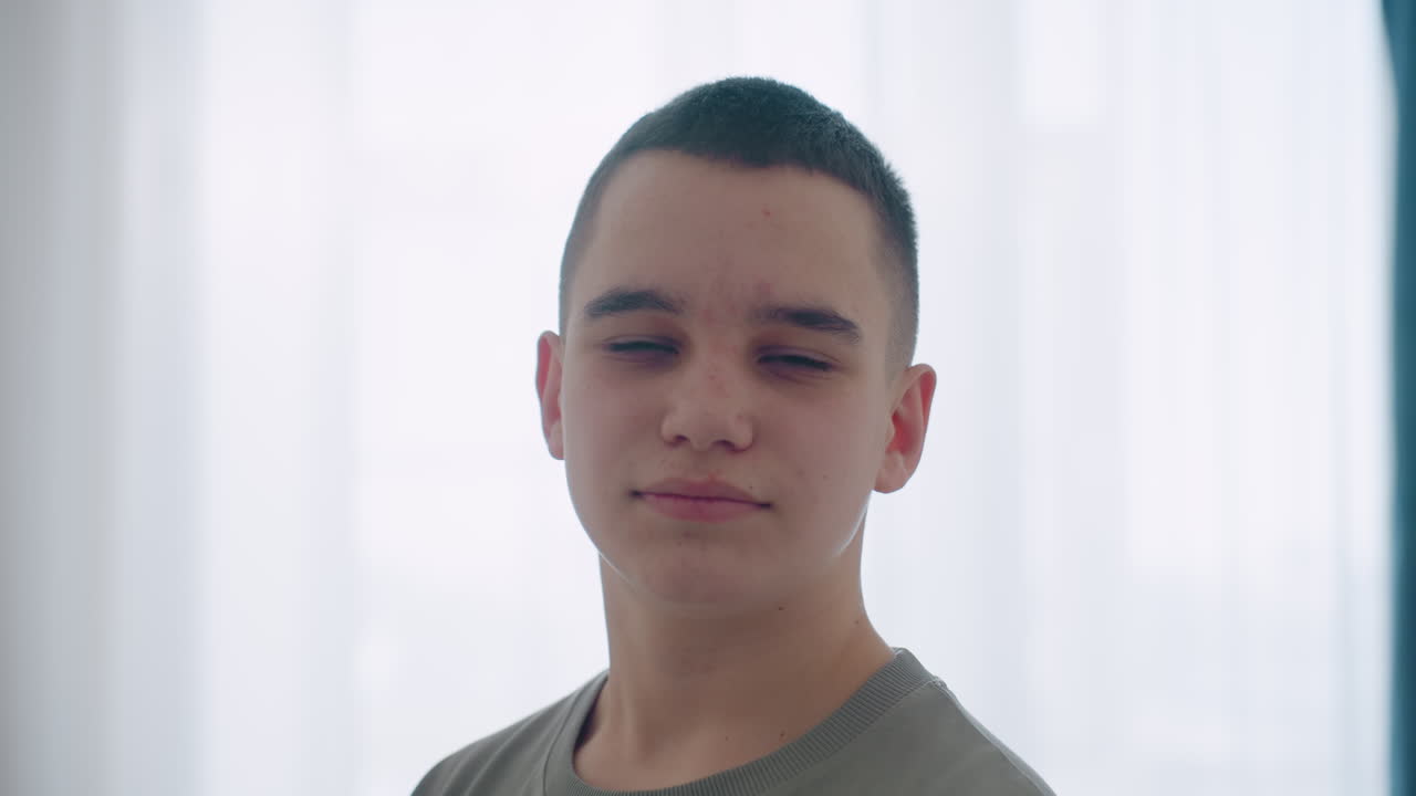 Cute boy turns around with gentle smile inside bright room, standing calmly in casual t shirt, natural light highlighting profile as he shows youthful innocence