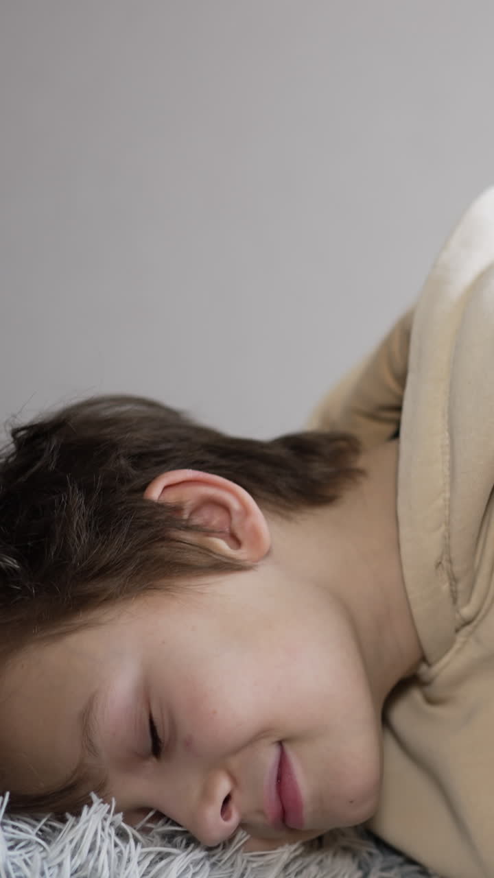 Handsome brunet boy holds a soft fluffy pillow in his hands. Teenager bends to put his head on the pillow. Close up. Light backdrop. Vertical video