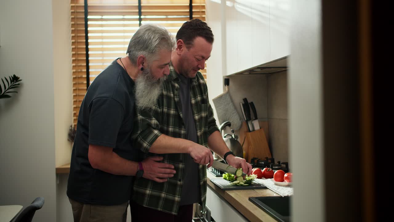 Happy middle-aged brunette man with stubble in a green shirt prepares a salad of tomatoes and cucumbers while his elderly boyfriend with gray hair and a lush beard hugs him from behind in the kitchen in the morning