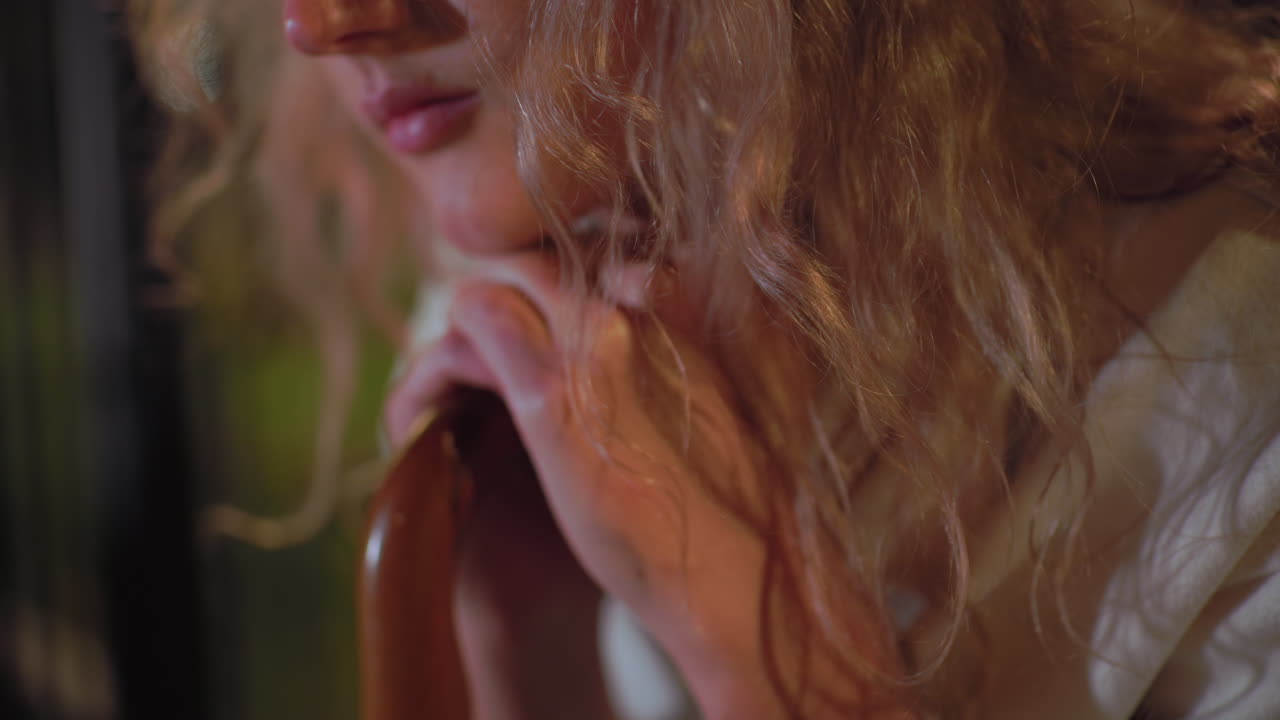 Close up of young lady with curly hair wearing white coat gently resting hands and chin on umbrella handle, appearing calm and peaceful under soft night lights with blurred outdoor background