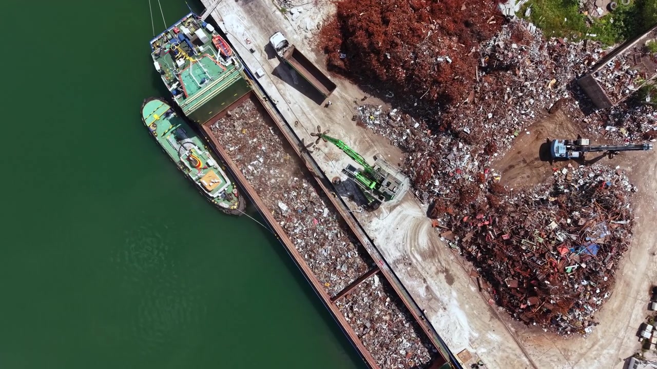 Operai che lavorano rottami metallici in un impianto di riciclaggio vicino all'acqua, in una giornata di sole, in una trafficata zona industriale.