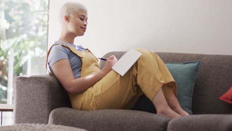 happy biracial woman taking notes and sitting on couch in slow motion