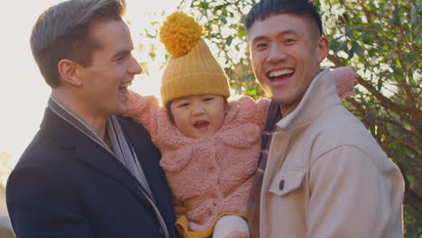 Portrait-Of-Family-With-Two-Dads-On-Walk-In-Winter-Countryside-Carrying-Daughter