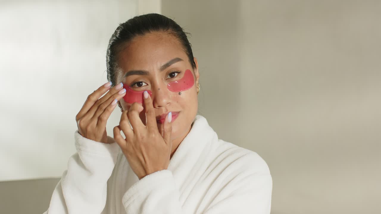 Once red gel under-eye patches applied, Asian woman in robe tapping, swiping at vanity for adhesion