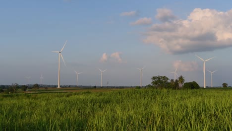 Wind-Turbines-shooting-out-of-a-Farmland-while-the-Sun-is-setting-as-Trucks-Pass-by-making-Clouds-of-Dust,-clean-alternative-energy-in-Thailand-and-mainland-Southeast-Asia