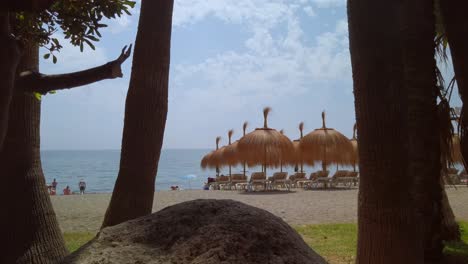 gimbal shot of tropical marbella beach with tikki umbrellas in background and rock and palm trees in forground