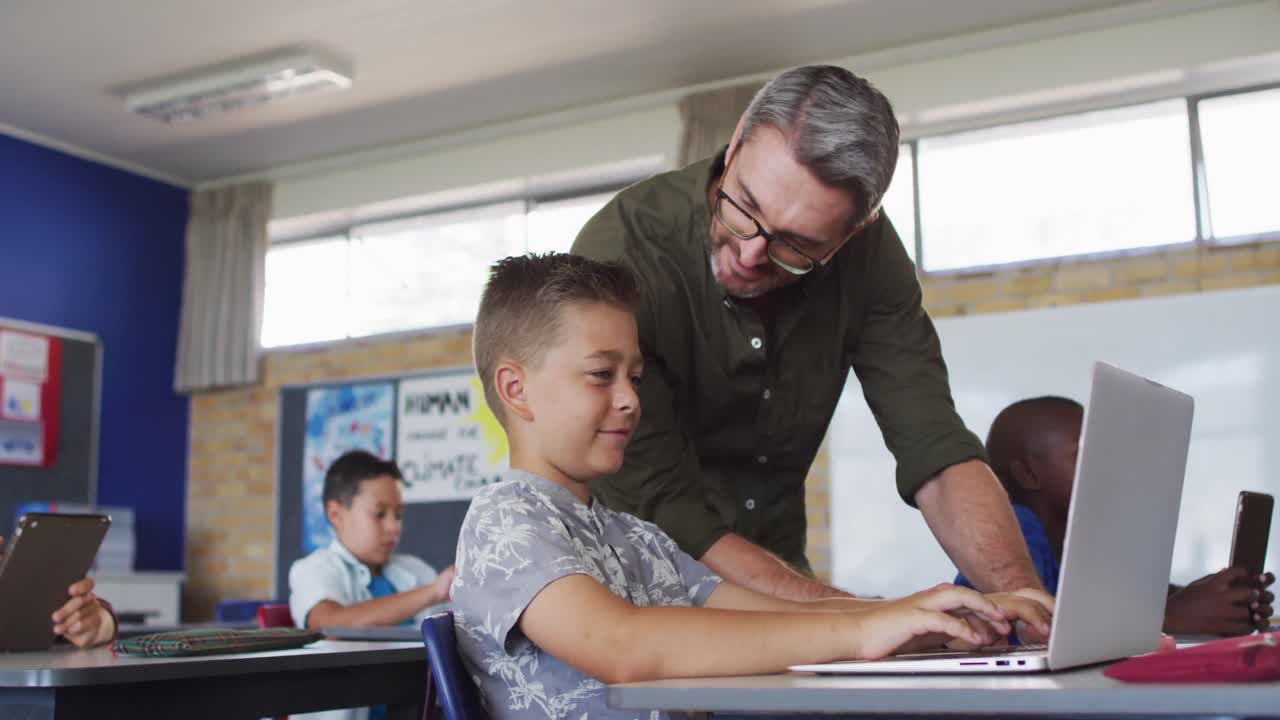 un maestro masculino diverso ayudando a un escolar sentado en el aula usando una computadora portátil