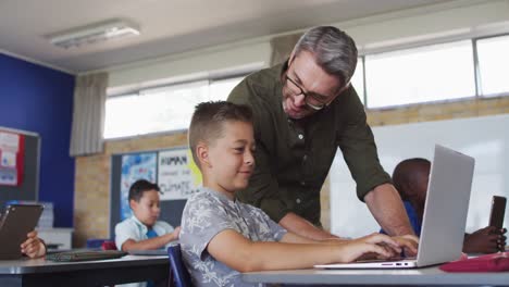 Diverse-male-teacher-helping-a-schoolboy-sitting-in-classroom-using-laptop