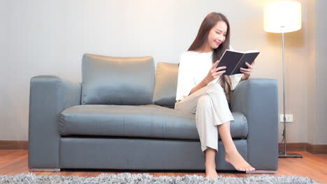 a young woman sits on a couch under a floor lamp reads a book