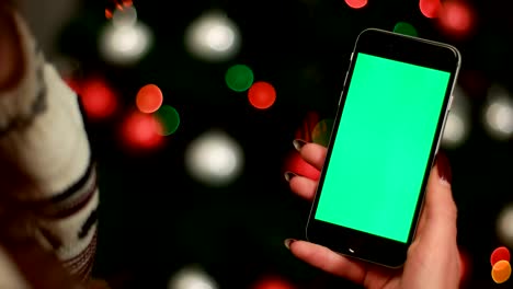 woman hand using smart phone with green screen on garlands bokeh from christmas tree background. close up. chroma key.