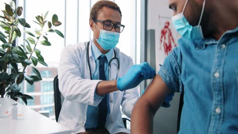 Close-Up-Of-Young-Happy-Hindu-Man-Patient-Receiving-Coronavirus-Vaccine-In-Hospital-Cabinet,-Handsome-Male-Physician-Specialist-In-Medical-Mask-Injecting-Patient,-Vaccination-Concept