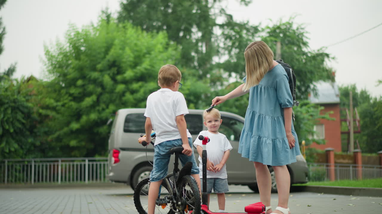 엄마는 만족해 보이는 어린 아들의 머리카락을 부드럽게 고, 그의 큰 형제는 자전거, 스쿠터, 주차된 차를 들고 근처에 서 있습니다.