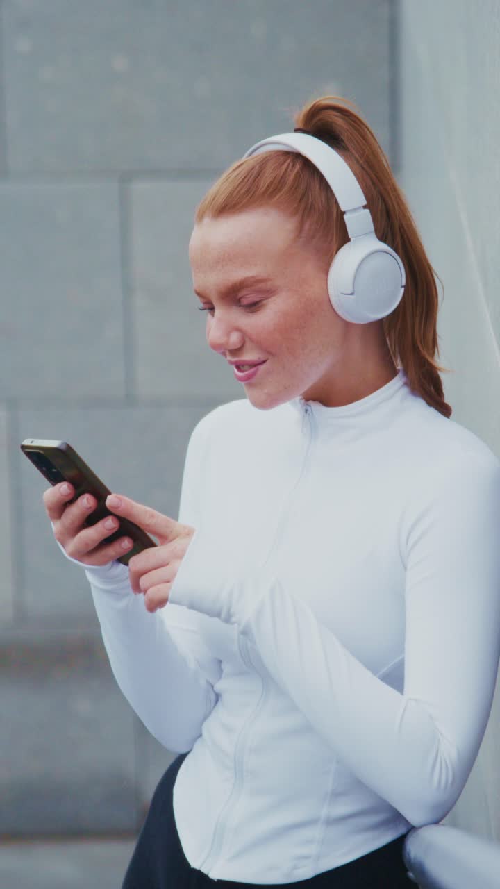 Young woman in white athletic wear enjoys music with headphones while using her smartphone, showcasing a moment of relaxation and modern lifestyle in a stylish urban setting