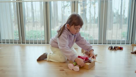 curious toddler girl playing with toys in an indoor play area, building and having fun with dolls on blocks, sideview medium static, slow motion