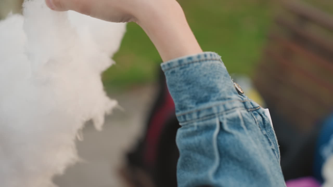Giovane padre caucasico offre zucchero filato, gita in famiglia su una panchina del parco, catturando un tenero momento di genitorialità e uno sguardo affettuoso verso il neonato che indossa berretto e giacca di jeans, con uno zaino sfocato.