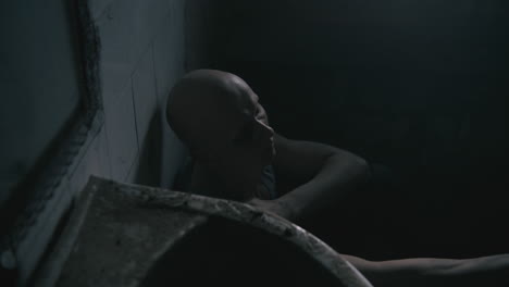 pale bald skinny man sits next to the wall in the creepy old bathroom at night