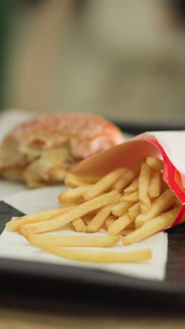 un primer plano de papas fritas, una hamburguesa y una taza de café en una bandeja, la mano de la mujer alcanza las papas frites, toma un bocado y toma su café, todo mientras un suave efecto de iluminación bokeh hace sonar en el fondo borroso