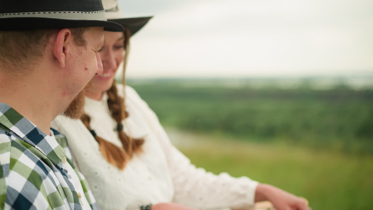 잔디에 있는 커플의 클로즈업, 검은 모자와 카레드 셔츠를 입은 남자, 그리고  드레스를 입은 그의 파트너. 그녀는 그에게 페인팅 브러쉬 세트를 건네주면서 따뜻하게 웃는다.