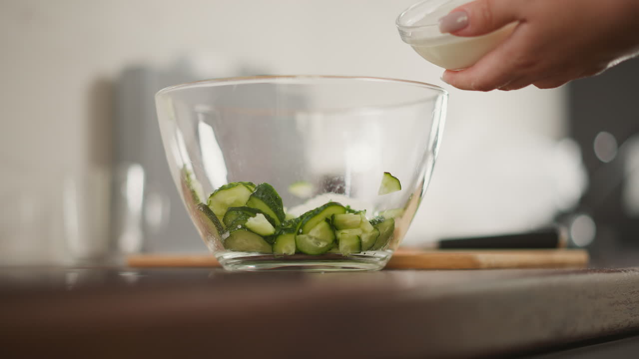 close up of glass container filled with freshly sliced cucumber on wooden counter while chef pours salad cream into bowl in kitchen with blurred background