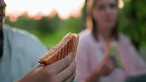 partial view of people enjoying sandwiches, slightly blurred background capturing a relaxed outdoor atmosphere, focus on the sandwich being held, with green nature and soft lighting in the distance