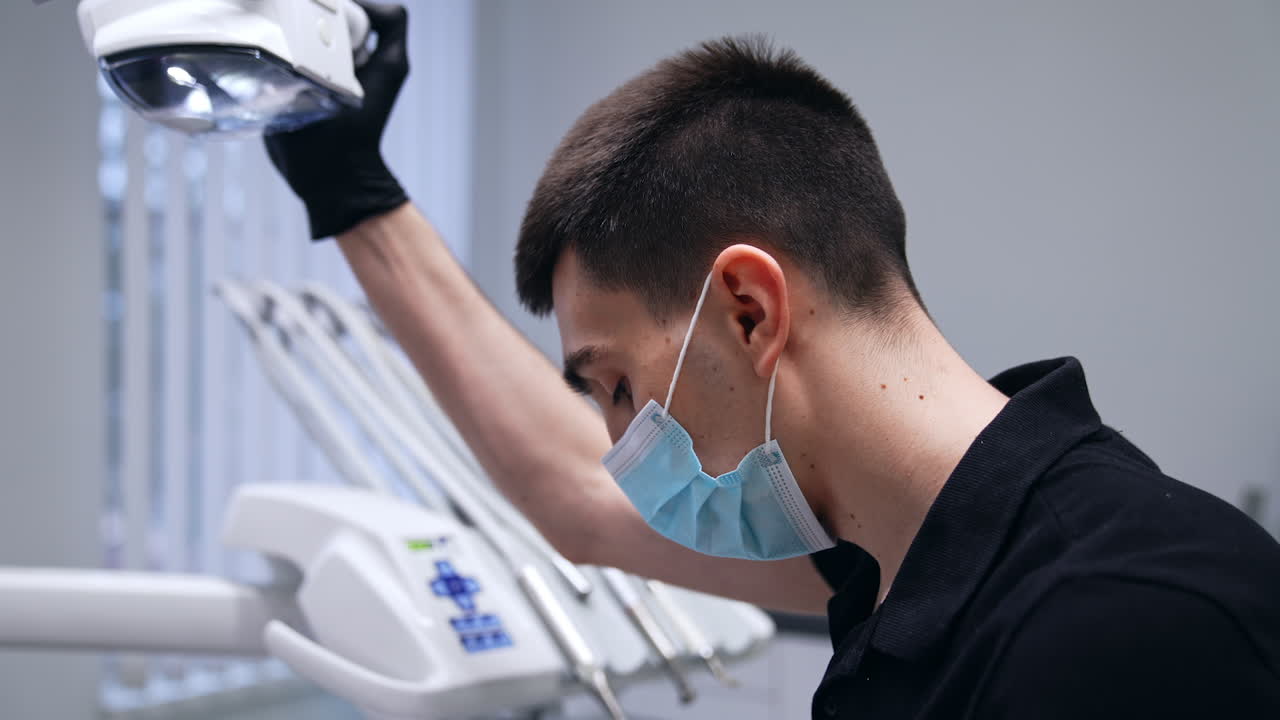 Young male brunet dentist in mask working in the cabinet. Doctor fixing the lamp position and takes the drill machine. Female nurse at foreground.