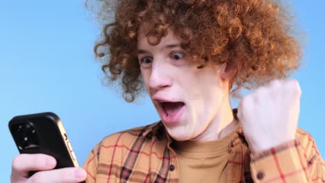 cute young happy student on blue background looking at phone cute young happy student on blue background looking at phone curly redhaired guy is crazy happy internet search technologies