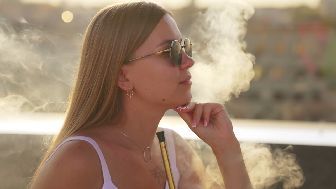 Leisure time in a café outdoors in summer. Lady in sunglasses and white dress smoking hookah. Side view. Close up.