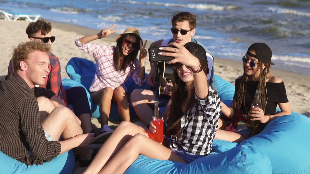 Young hipster girl recording a video or taking selfie of group of friends sitting on easychairs on the beach, playing guitar and singing on a summer evening. Slowmotion shot