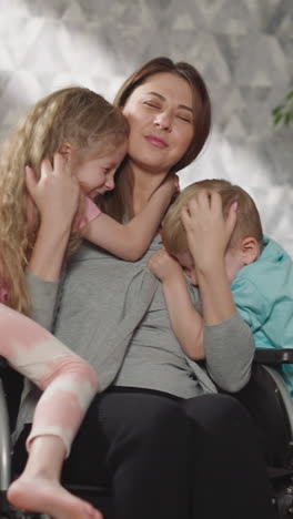 loving children come to hug mother sitting in wheelchair in living room. woman with spinal cord injury enjoys spending time with little son and daughter