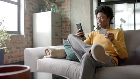 happy african american man sitting on sofa using smartphone for online shopping at home, slow motion