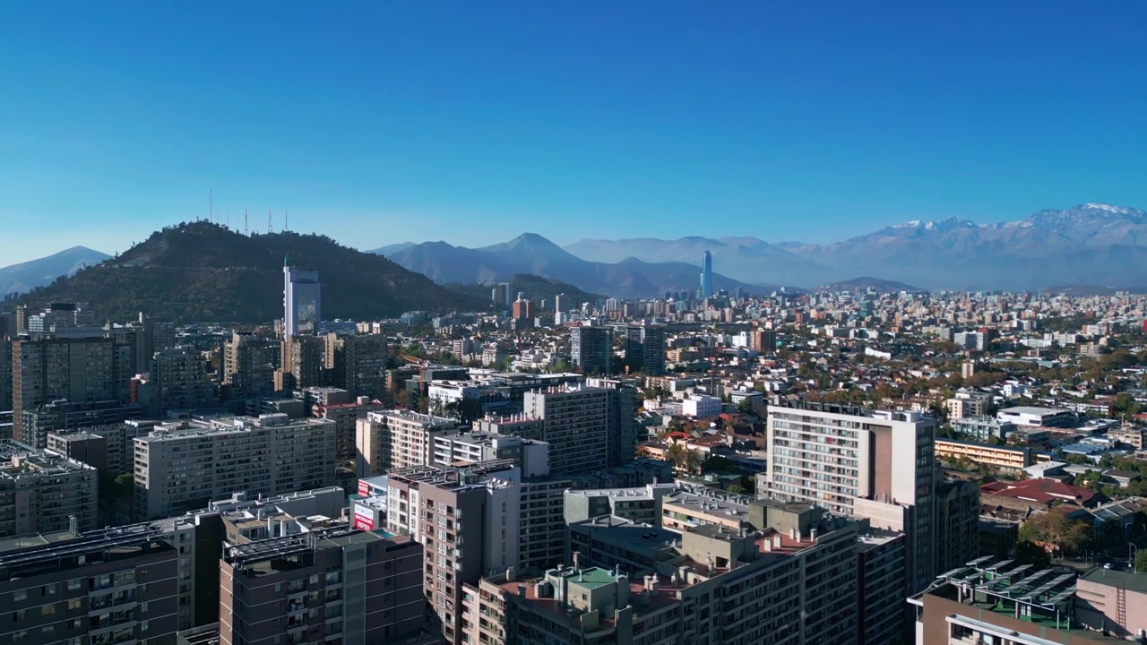 Lenta ripresa ascendente sopra un denso quartiere residenziale che rivela lo skyline di Santiago, il Cerro San Cristóbal e la cordigliera delle Ande innevate sotto un cielo azzurro limpido. Santiago, Cile.