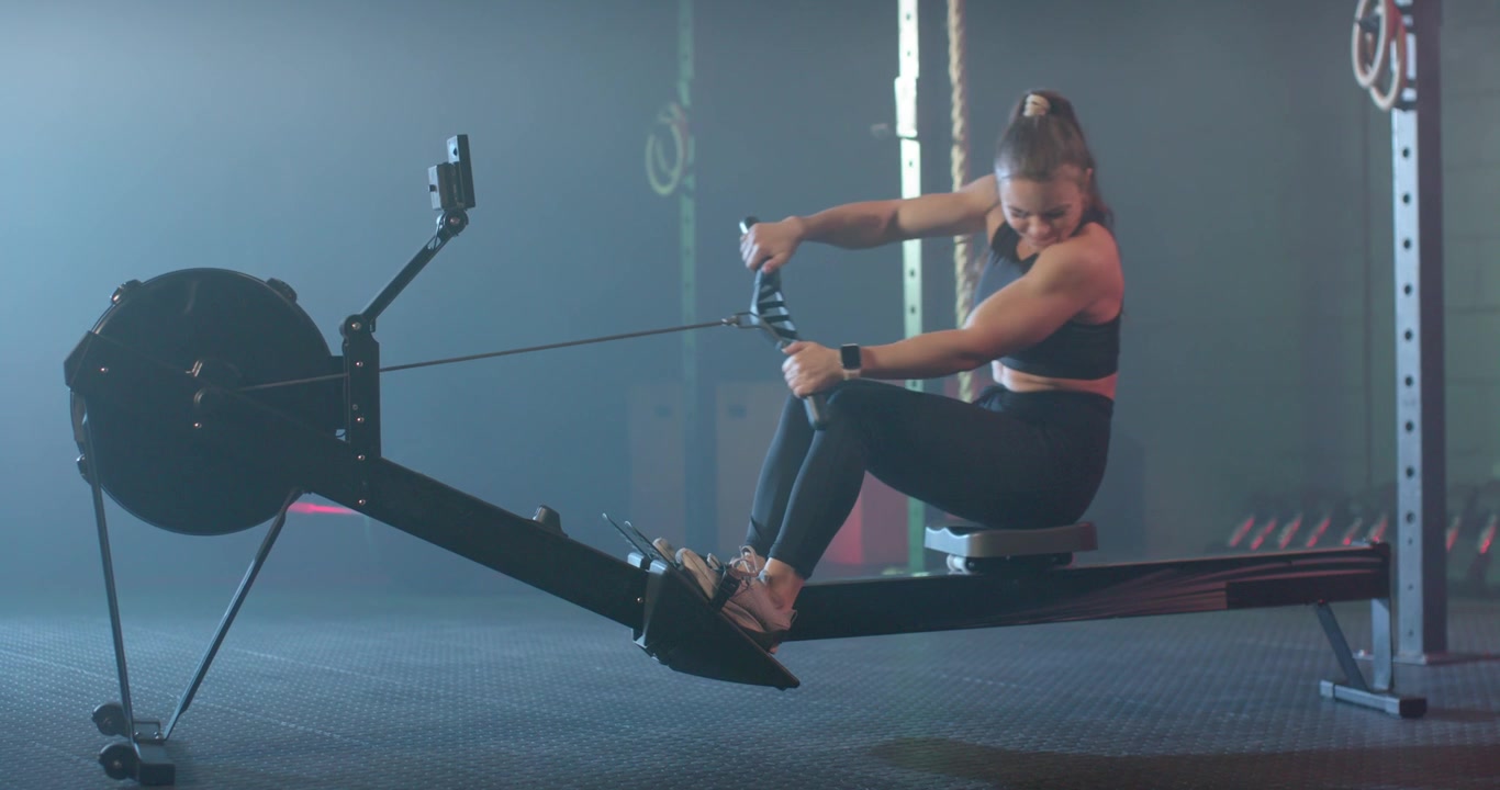 Woman in black bra tights using rowing machine in gym, driving feet pulling handle tracking monitor
