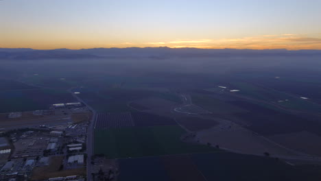 Vista-Aérea-De-Los-Campos-Que-Rodean-La-Pequeña-Ciudad-Con-El-Sol-Saliendo-Por-Encima-De-Las-Montañas