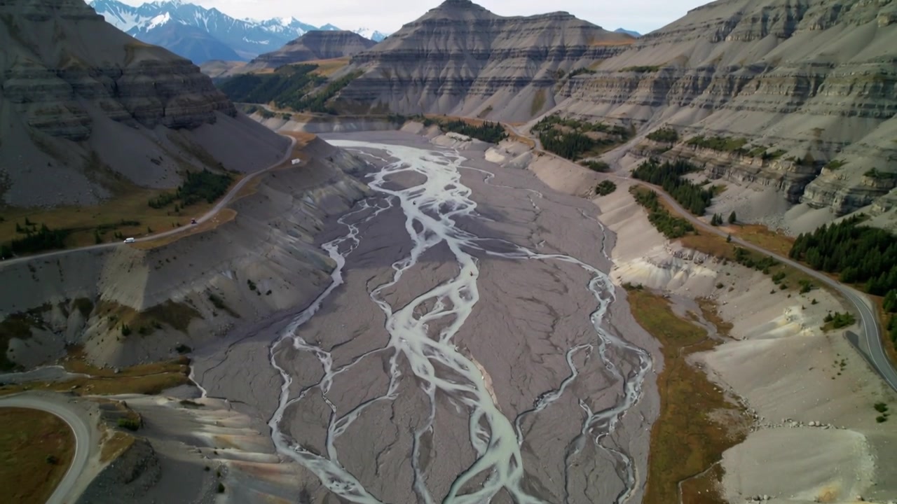 La telecamera inizia una panoramica, un drone scende su una valle fluviale a canali intrecciati, rivelando banchi di ghiaia e una strada sterrata.