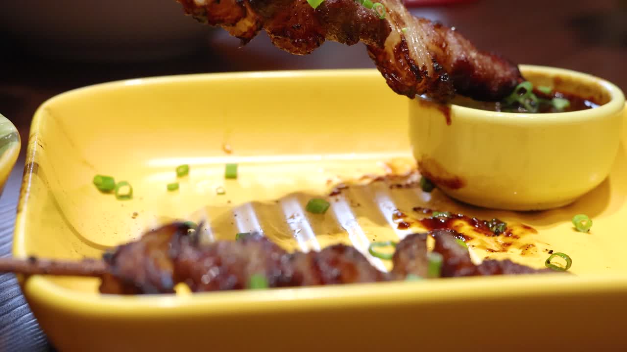 Juicy grilled beef skewers are lifted and dipped into a savory sauce on a yellow plate under warm indoor lighting, highlighting Thai cuisine presentation