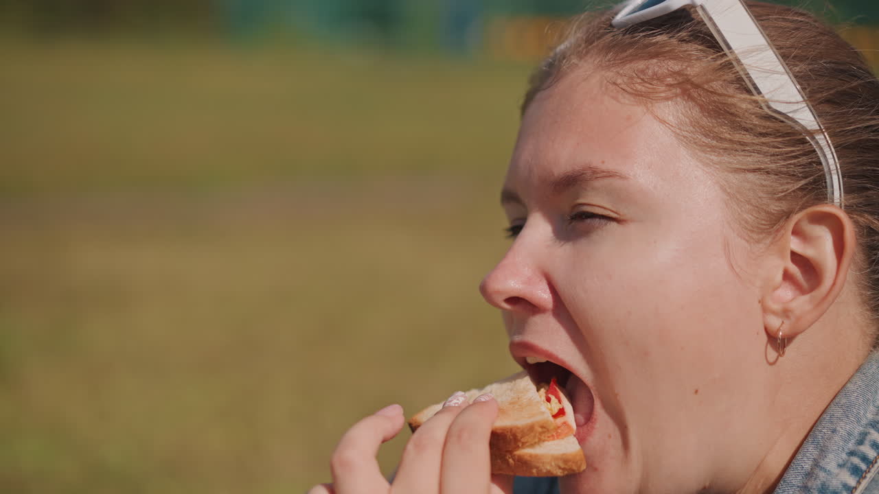 Young woman eating sandwich outdoors, sunlit profile shot captures casual denim jacket, grassy field backdrop, joyful bite with crumbs, slow savor and relaxed weekend picnic vibe, candid lifestyle