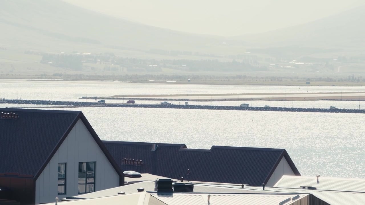 A high-angle view over rooftops in Akureyri, Iceland, shows cars driving on a causeway across a sunny, glistening fjord with hazy mountains in the background.