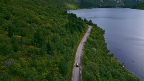 downwards facing shot of golden car driving along the scenic road on the coast through the green forests in northern europe norway lofoten with view towards the blue ocean and high dramatic mountains