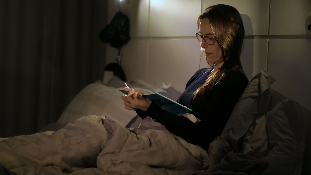 Adult woman puts on her glasses and reads a novel in bed. Moody night lighting, Medium shot
