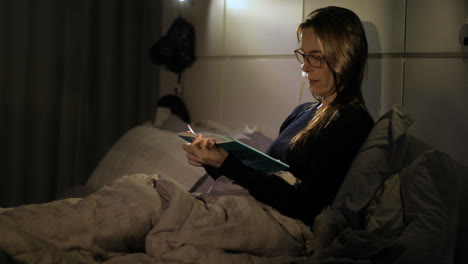 adult woman puts on her glasses and reads a novel in bed. moody night lighting, medium shot