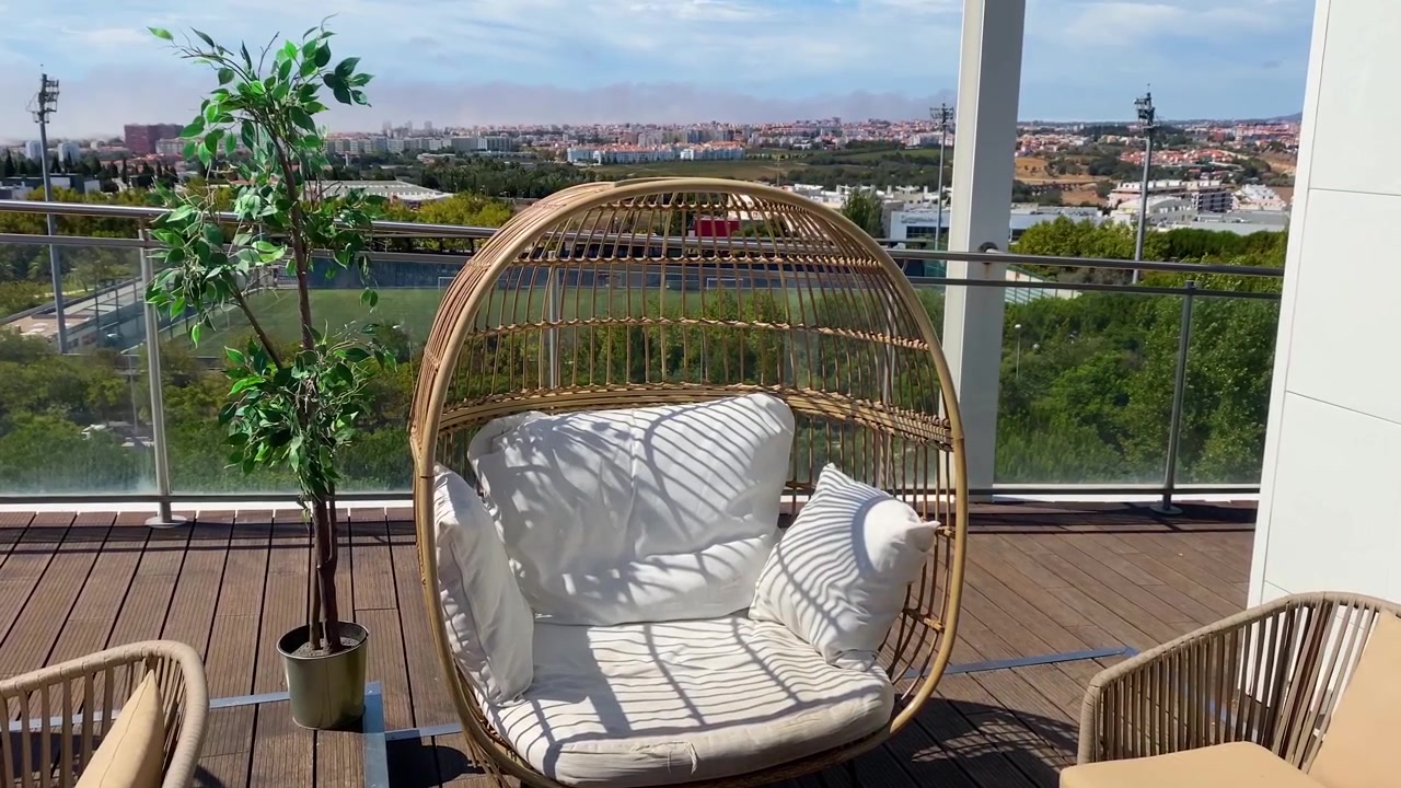 Balcone moderno con una comoda sedia in rattan e una pianta in vaso, bagnato dalla calda luce del sole. Ambiente perfetto per il relax, il tempo libero o per godersi la vista urbana in serena comodità.