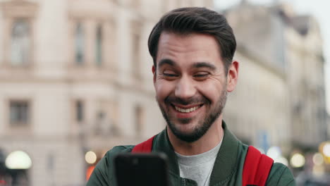 Vista-Cercana-Del-Joven-Caucásico-Con-Mochila-Enviando-Mensajes-De-Texto-En-Su-Teléfono-Inteligente-Alegremente-En-La-Calle