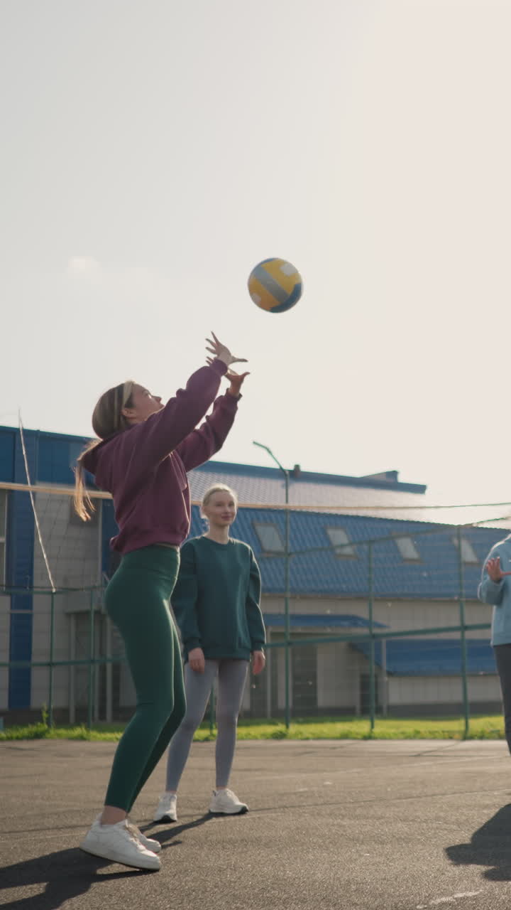 배구 코치와 함께 야외에서 훈련하는 배구 초보자, 다른 선수들로 둘러싸여, 녹색 바 울타리 뒤에서 빛으로 빛나는 코트에서 건물을 배경으로 고 있습니다.