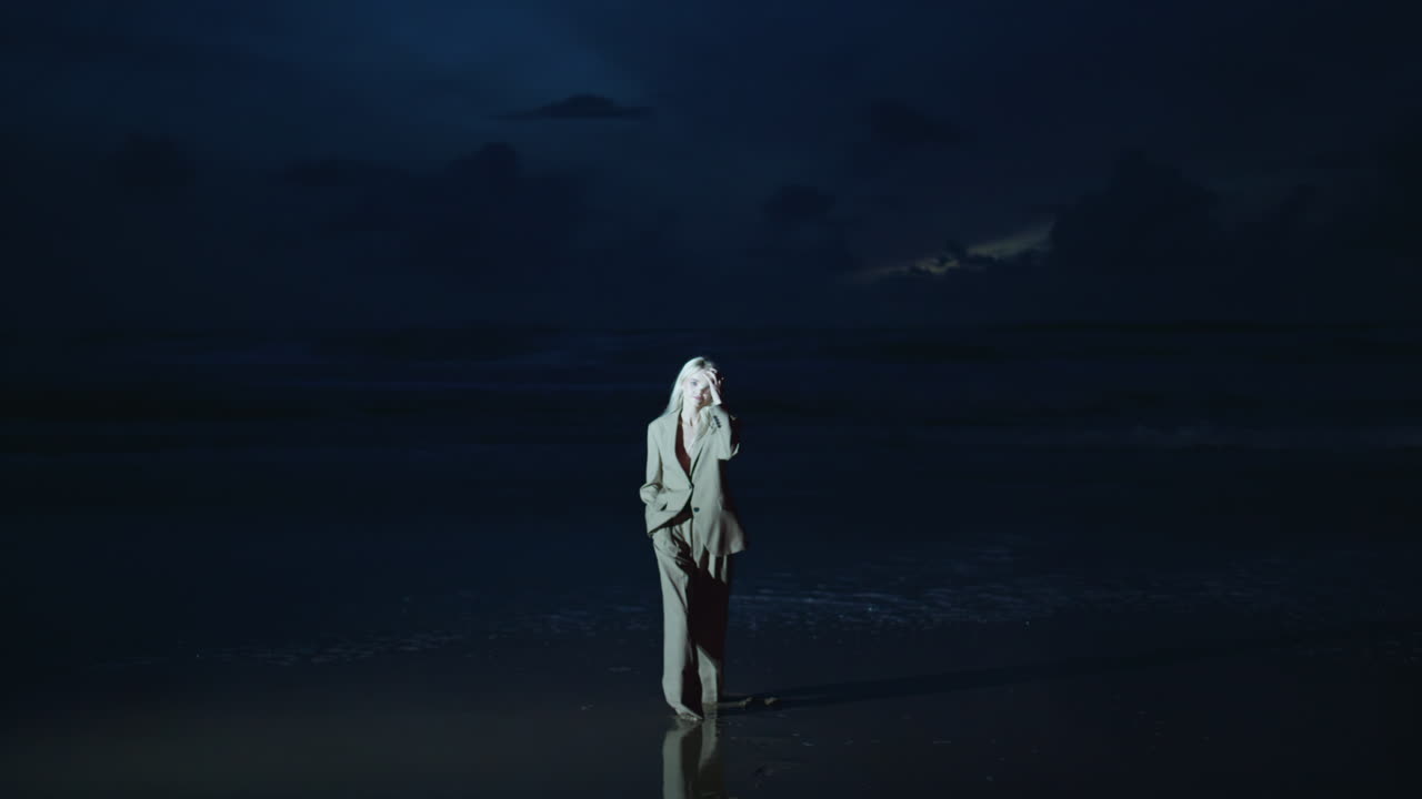 Işıklandırılmış gece sahilinde modellik yapan kız. Karanlık sahilde duran takım elbiseli rahat kadın.
